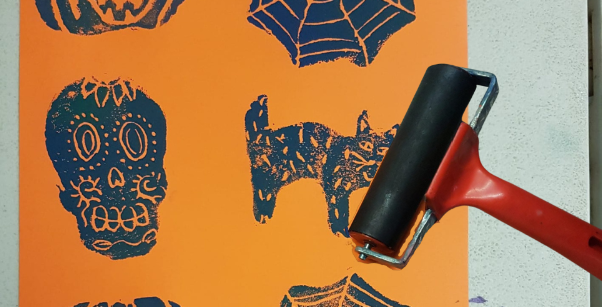 An image of an orange piece of paper with black halloween symbols such as a skull and a cat screenprinted on. There is also a screenprinting roller on the right side.