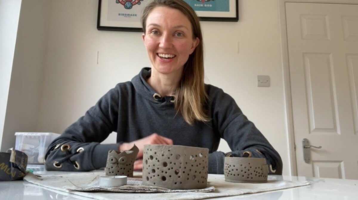 A female artist with long, dark blonde hair, wearing a grey sweatshirt, sits at a table with some handmade pottery - two cylindrical objects with hole markings and a small clay owl.