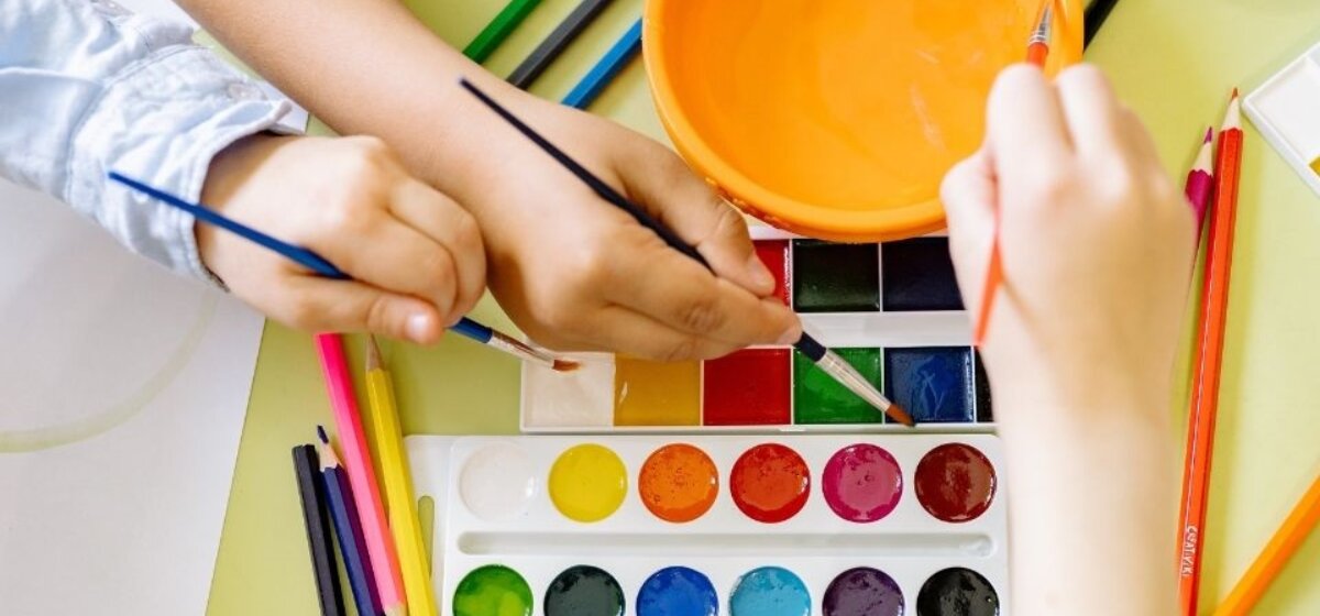 Children's hands over a paint palette with paint brushes and an orange bowl