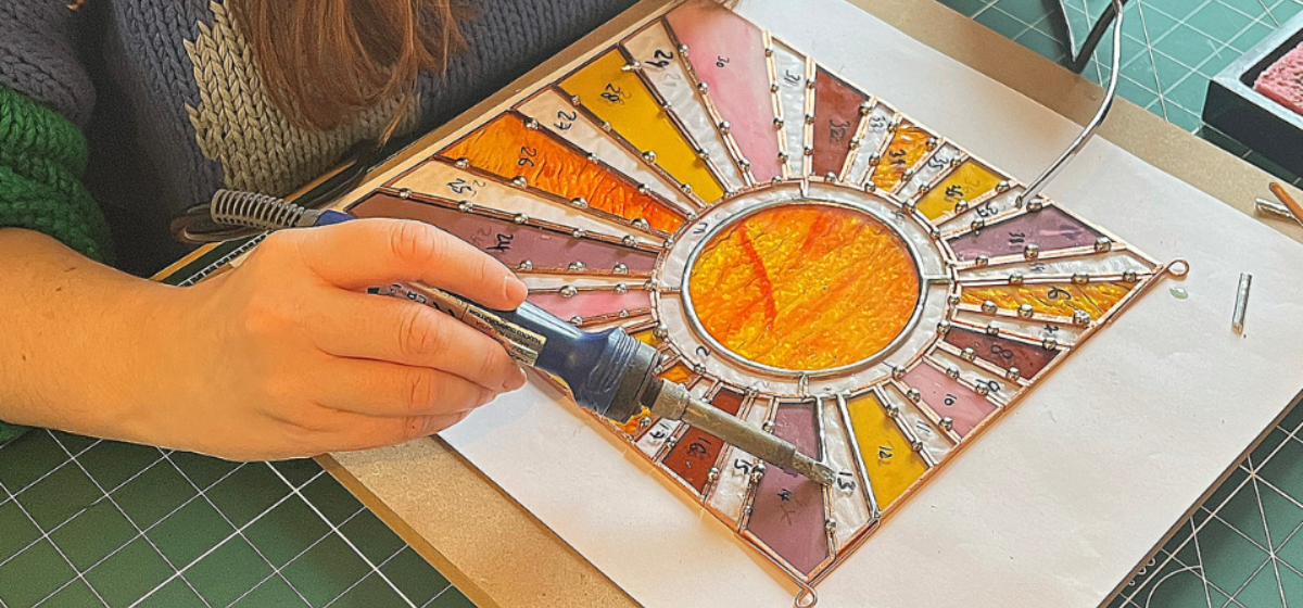 A person using a soldering iron to solder together glass pieces lined with copper wire. The glass create the image of a gleaming sun