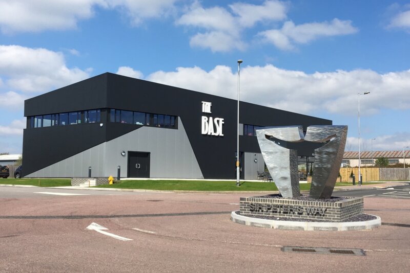 A distanced outside shot of The Base building. In the foreground is a round about with a monument on it. It's a sunny day.