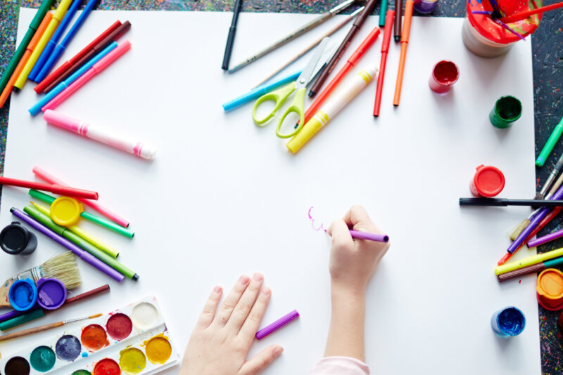The hands of a child drawing on white paper surrounded by art materials.