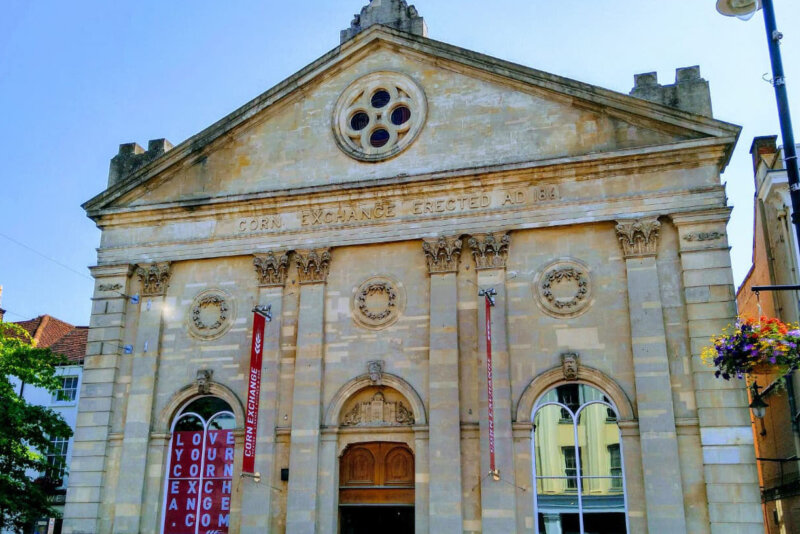 The front of the Corn Exchange building (opened in the 1800s, it is an old and grand building with two large windows at the front and a wooden door in the centre). Above the building there is a clear blue sky. There is a tree on the left with lots of green leaves and some hanging baskets are on a lamp post to the right of the building. The photo has been taken in the summertime.